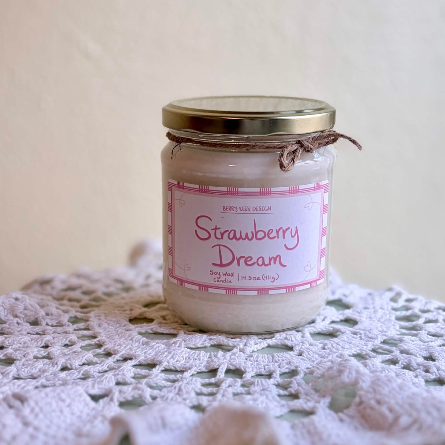 A glass jar candle labeled 'Strawberry Dream' sits on a white crocheted doily against a soft beige background.