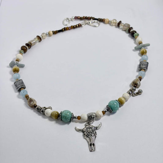 A beaded necklace with a silver-toned steer skull pendant, featuring light blue, white, brown, and gray beads, displayed on a white surface.