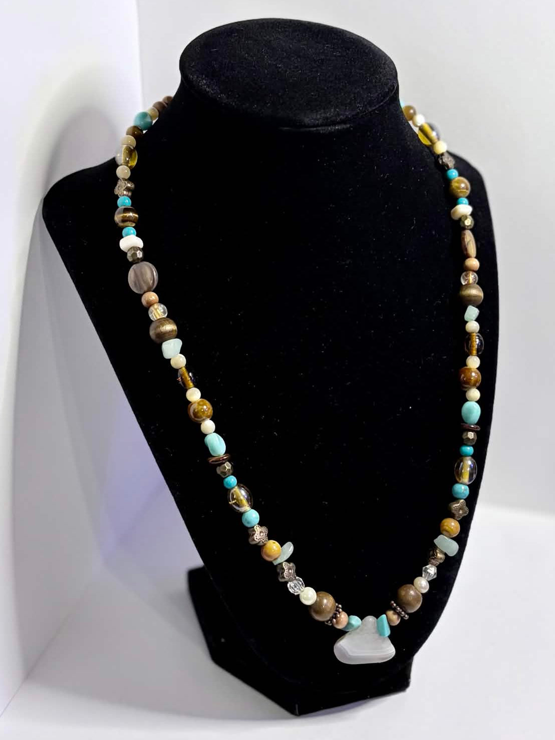 A beaded necklace displayed on a black velvet jewelry bust, set against a white photo backdrop. The necklace features small multicolored beads in shades of blue, green, brown, and gold, with a small white agate charm at the center.