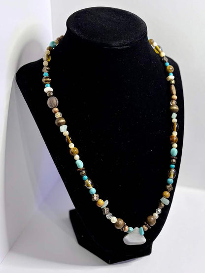 A beaded necklace displayed on a black velvet jewelry bust, set against a white photo backdrop. The necklace features small multicolored beads in shades of blue, green, brown, and gold, with a small white agate charm at the center.