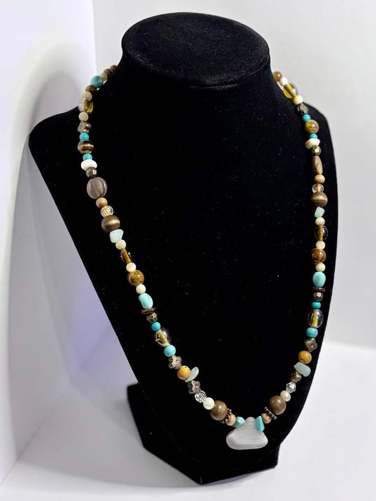 A beaded necklace displayed on a black velvet jewelry bust, set against a white photo backdrop. The necklace features small multicolored beads in shades of blue, green, brown, and gold, with a small white agate charm at the center.