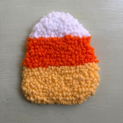 A punch needled coaster shaped like a candy corn, with white on top, orange in the middle, and pale yellow on the bottom, placed on a table.