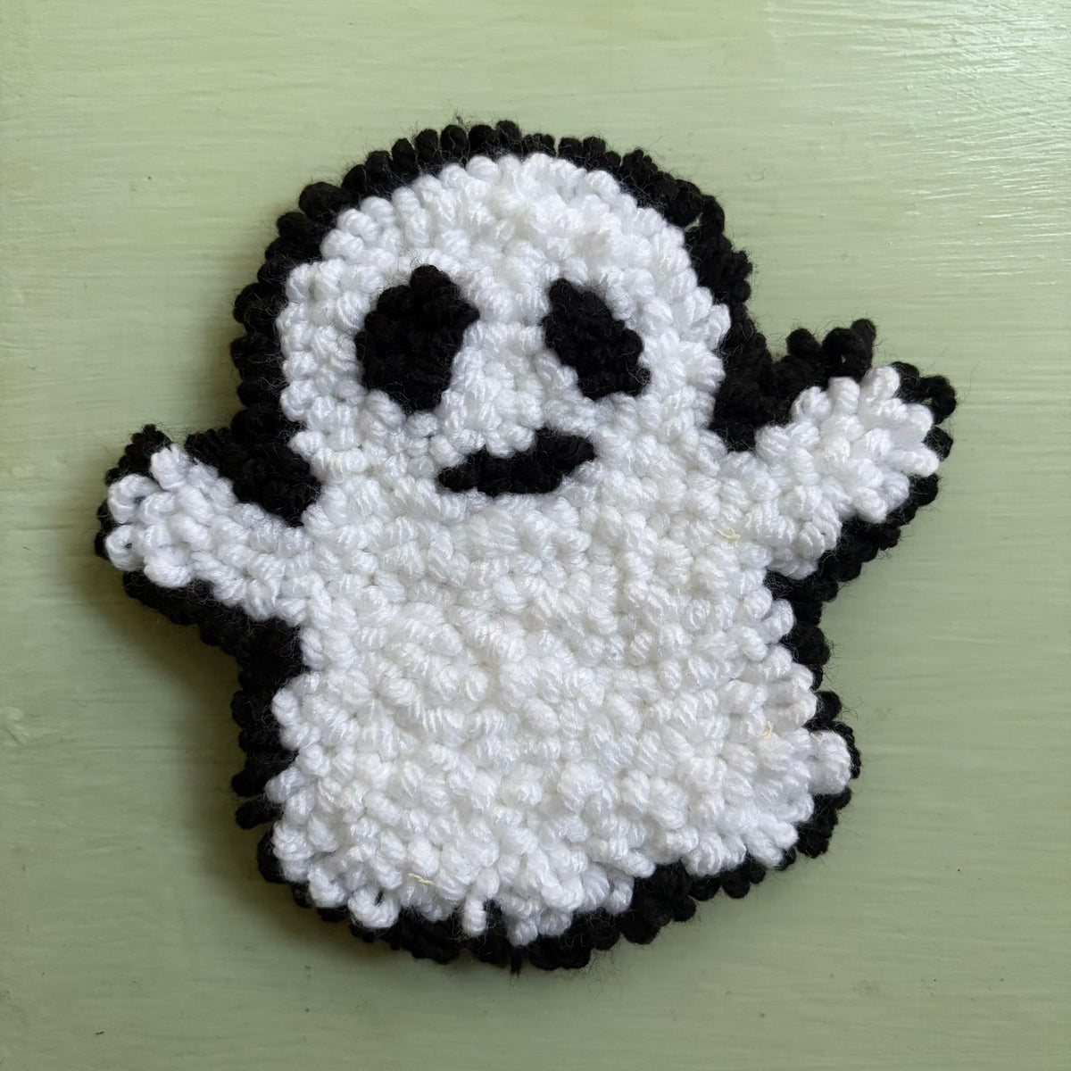 A handmade punch needle ghost mug coaster in white and black yarn, displayed on a table
