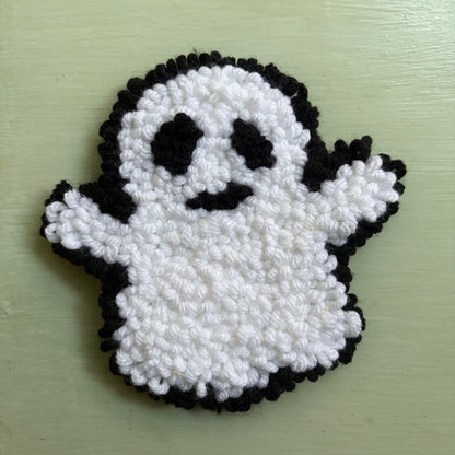 A handmade punch needle ghost mug coaster in white and black yarn, displayed on a table