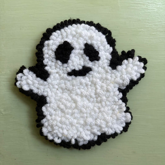 A handmade punch needle ghost mug coaster in white and black yarn, displayed on a table