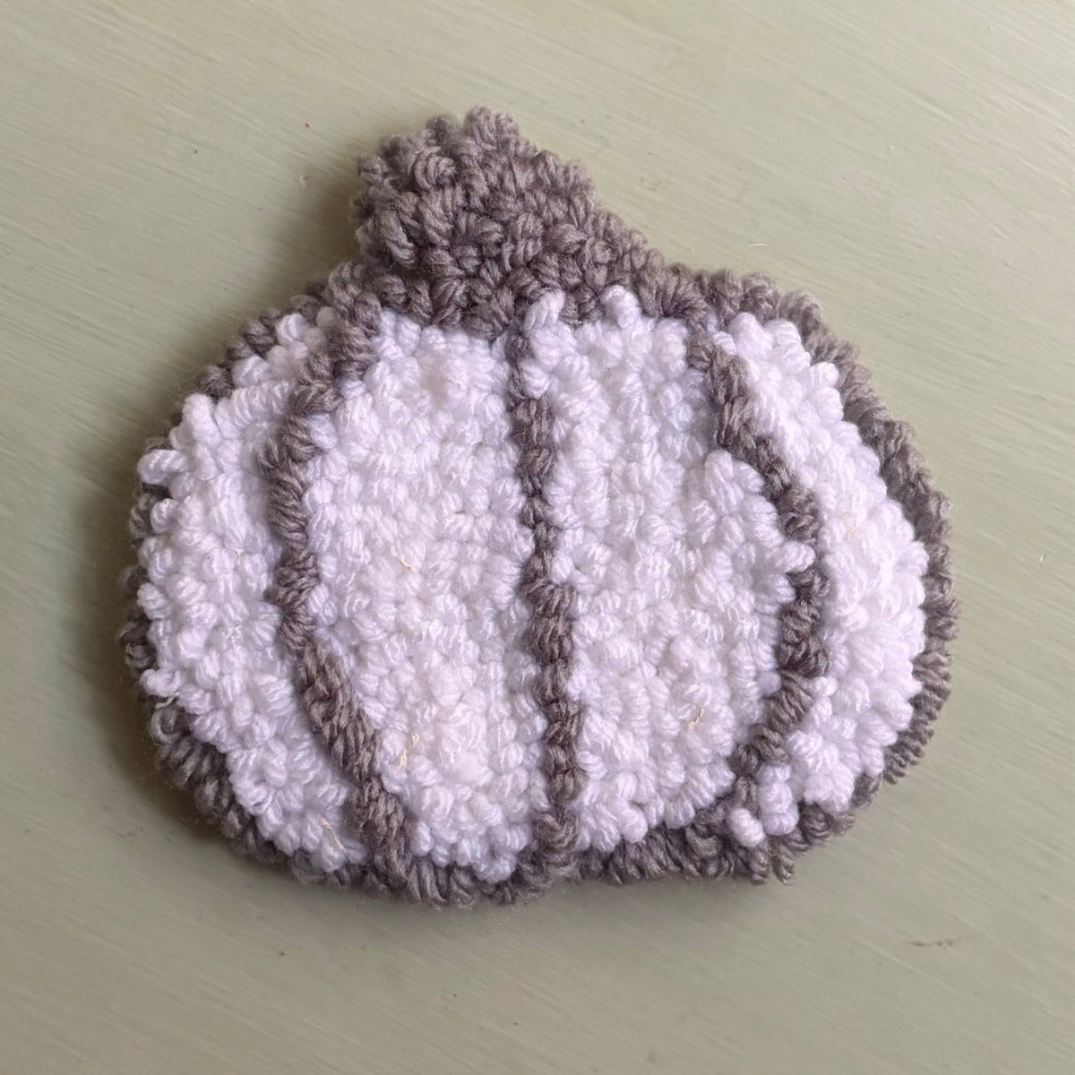 A punch needled coaster pumpkin-shaped white and gray, photographed on a table.