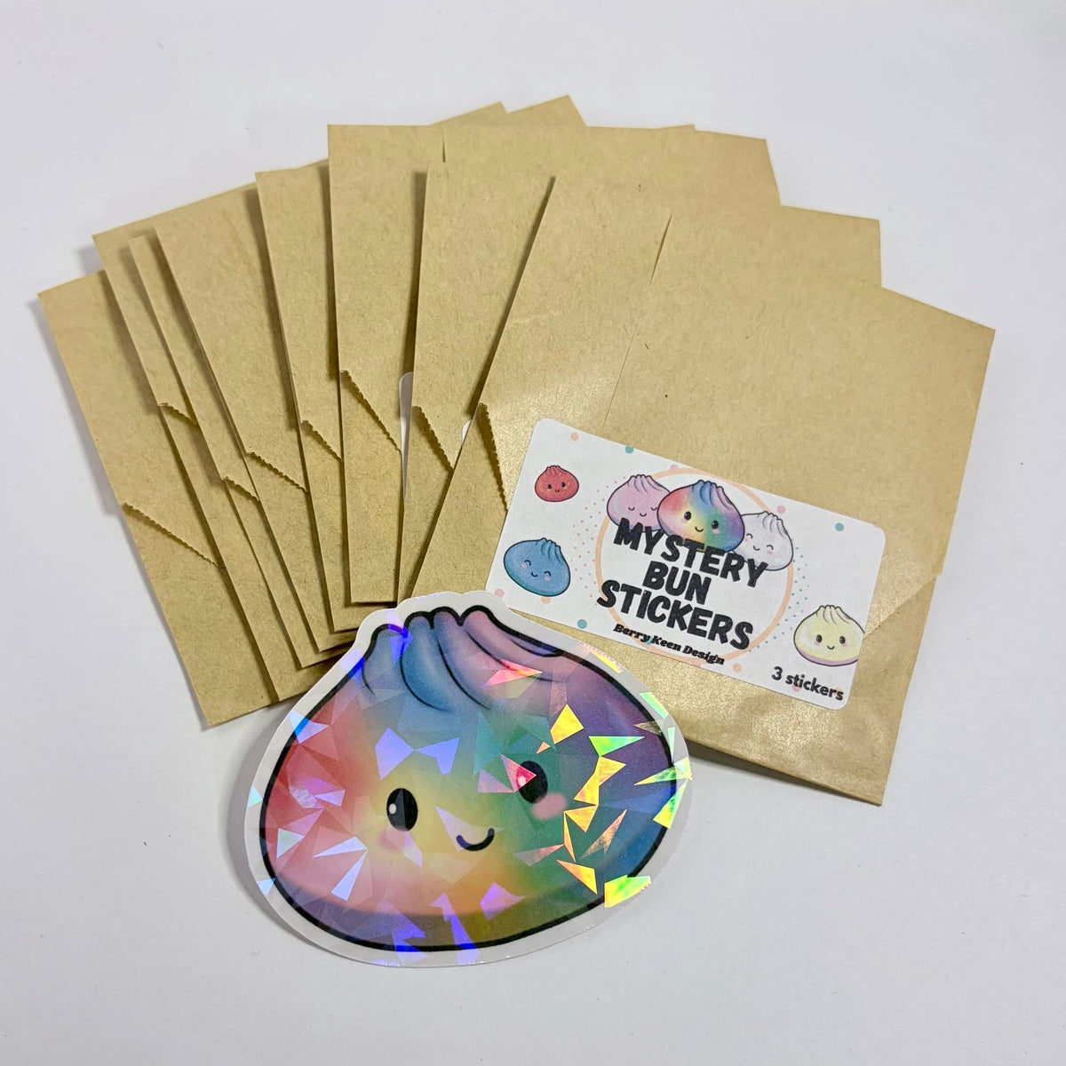 Brown kraft paper mystery envelopes with a 'Mystery Bun Stickers' label, alongside a single colorful sticker of a smiling bun with holographic foil accents, displayed on a white surface.