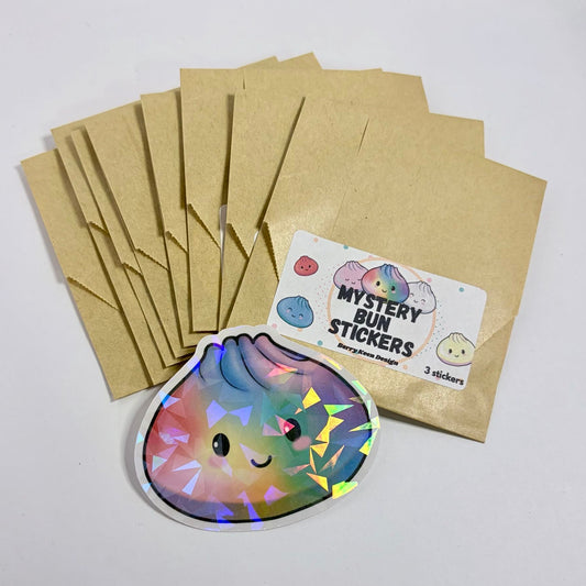 Brown kraft paper mystery envelopes with a 'Mystery Bun Stickers' label, alongside a single colorful sticker of a smiling bun with holographic foil accents, displayed on a white surface.