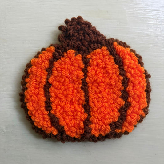 A punch needled pumpkin-shaped coaster in bright orange and dark brown yarn, photographed from above on a table