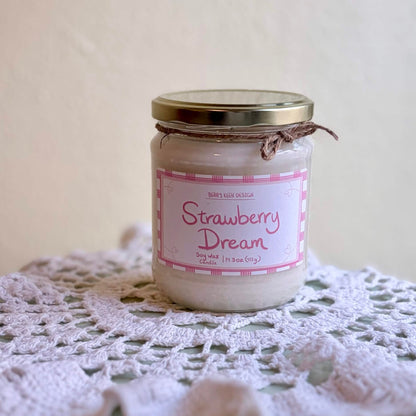A glass jar candle labeled 'Strawberry Dream' sits on a white crocheted doily against a soft beige background.