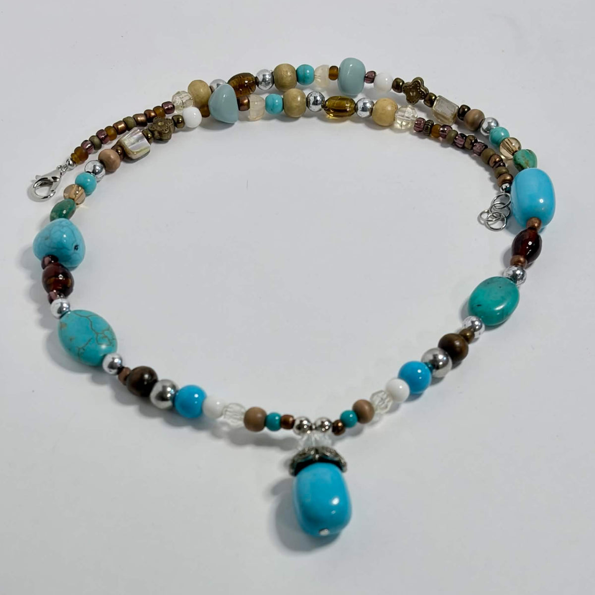 A handmade beaded necklace with turquoise, brown, white, and silver beads, featuring a large turquoise pendant, displayed on a white surface.