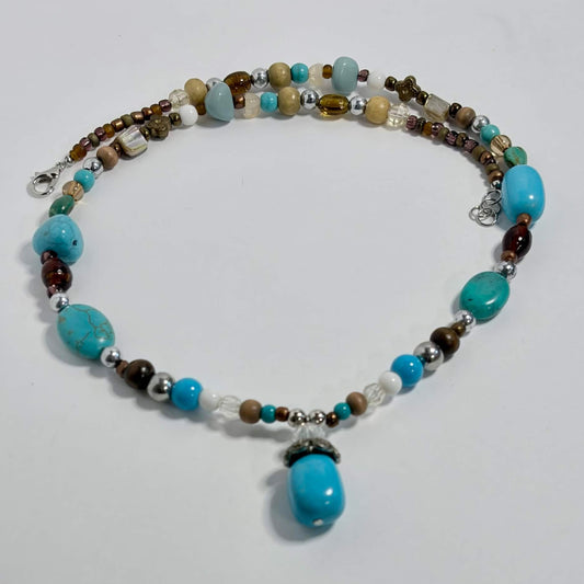 A handmade beaded necklace with turquoise, brown, white, and silver beads, featuring a large turquoise pendant, displayed on a white surface.