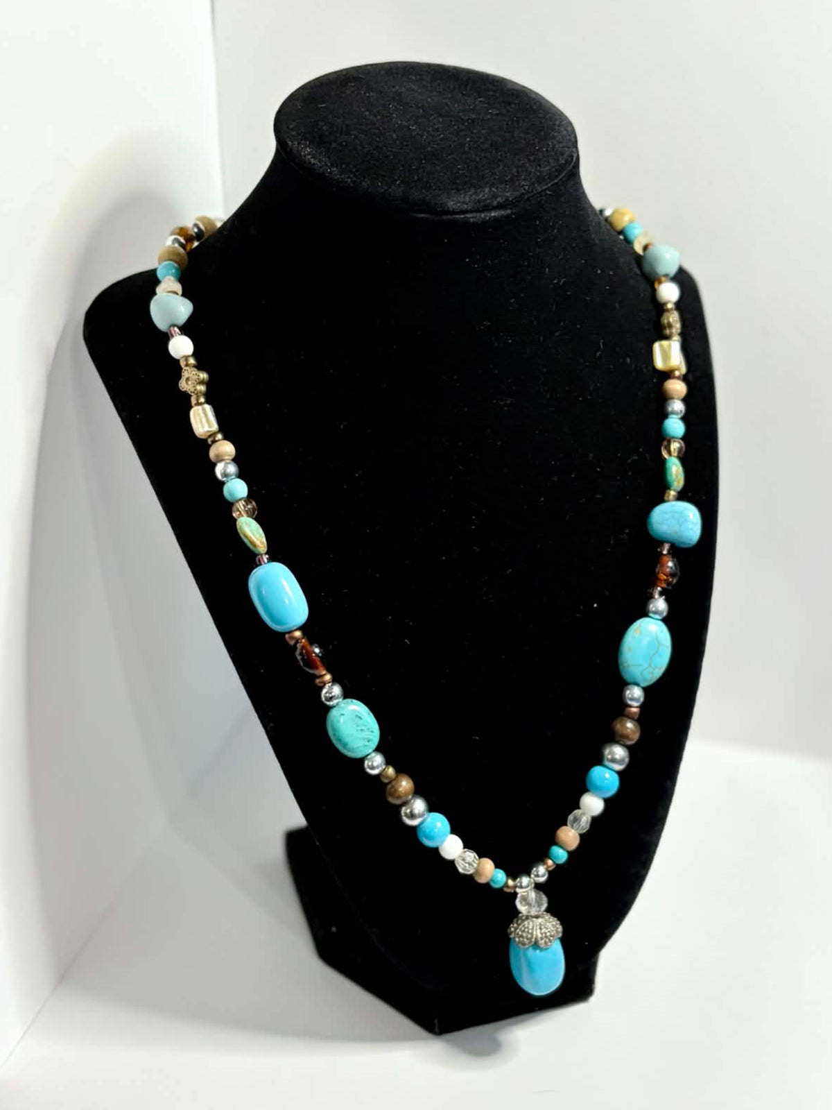 A beaded necklace with turquoise blue, white, and metallic beads displayed on a black jewelry bust, set against a white photo backdrop in a workshop environment.