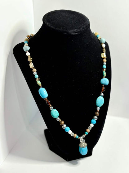 A beaded necklace with turquoise blue, white, and metallic beads displayed on a black jewelry bust, set against a white photo backdrop in a workshop environment.
