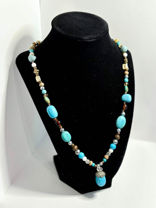 A beaded necklace with turquoise blue, white, and metallic beads displayed on a black jewelry bust, set against a white photo backdrop in a workshop environment.