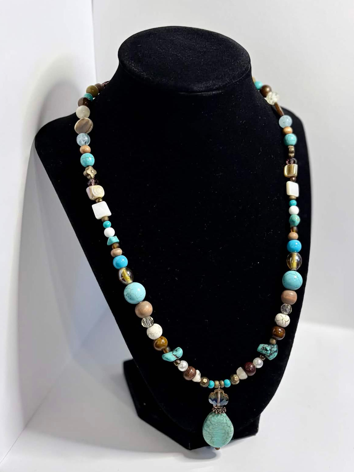 A beaded necklace with turquoise, white, and brown beads displayed on a black velvet jewelry bust, set against a white photo backdrop.