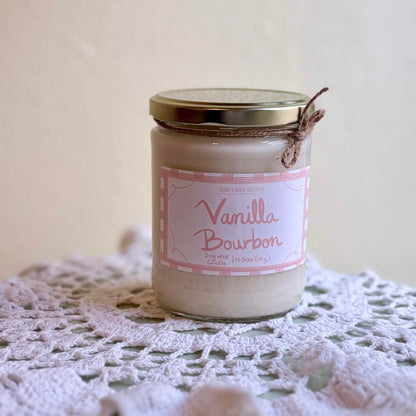 A glass jar candle labeled 'Vanilla Bourbon' sits on a white crocheted doily against a soft beige background.