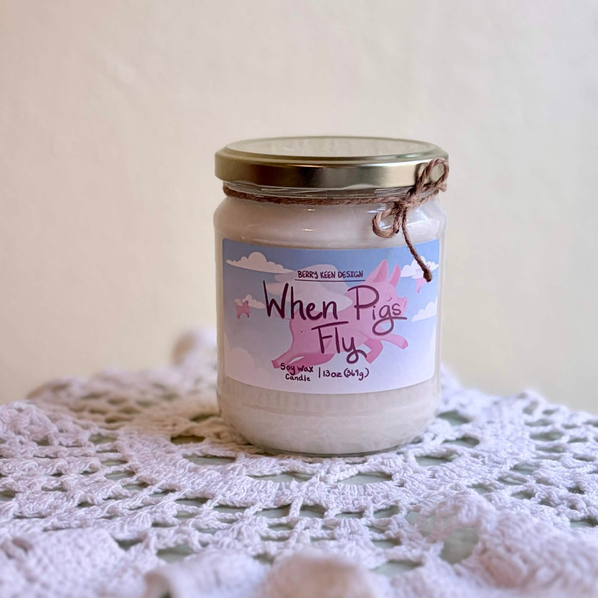 A jar candle labeled 'When Pig Fly' sits on a white crocheted doily against a soft beige background.