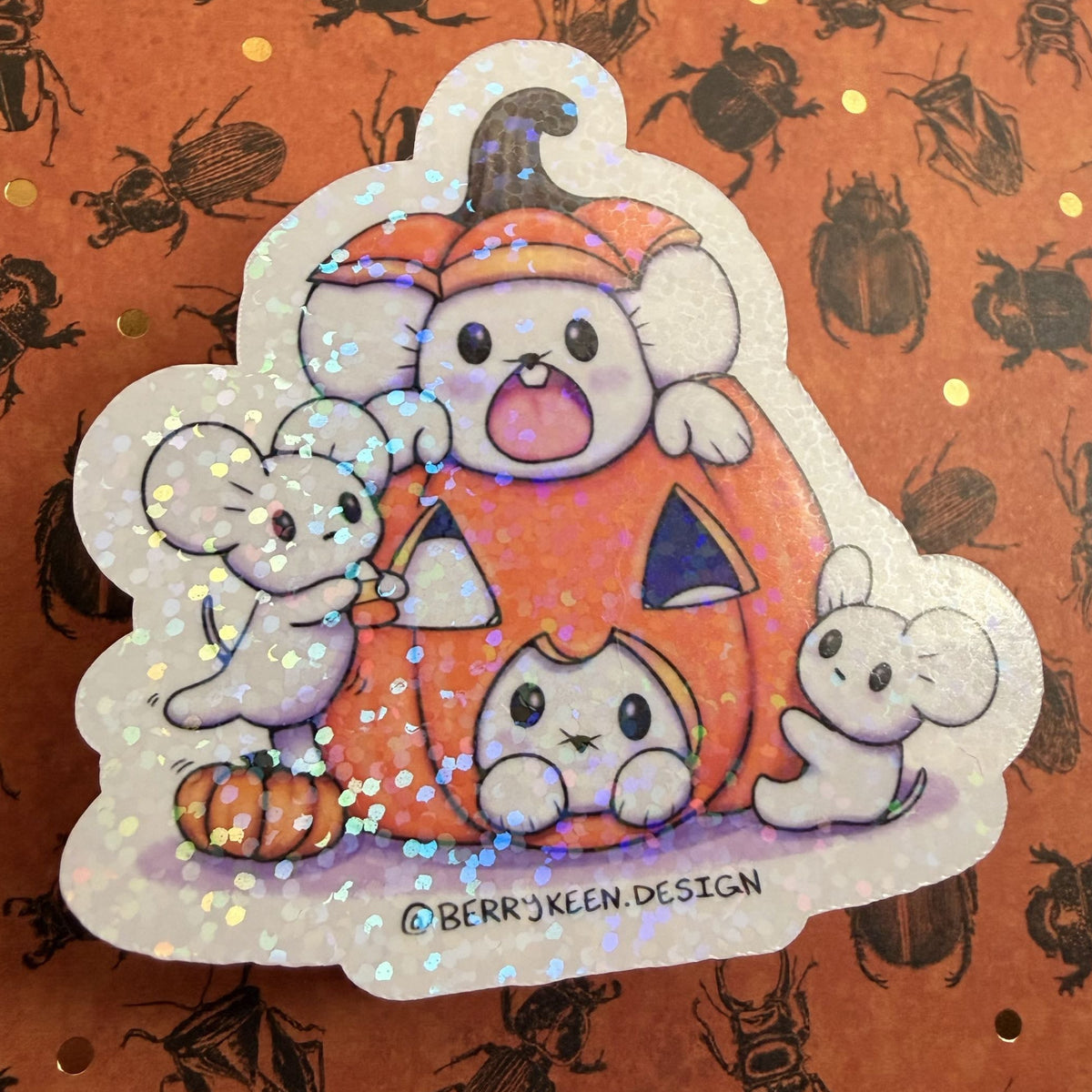 A die-cut sticker featuring three cute cartoon mice in a Halloween pumpkin, decorated with holographic glitter. The sticker is displayed on a orange background patterned with black beetle silhouettes.