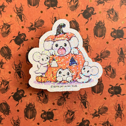 A die-cut sticker featuring three cute cartoon mice inside an orange pumpkin, placed on top of an orange background patterned with black beetles and small gold dots.