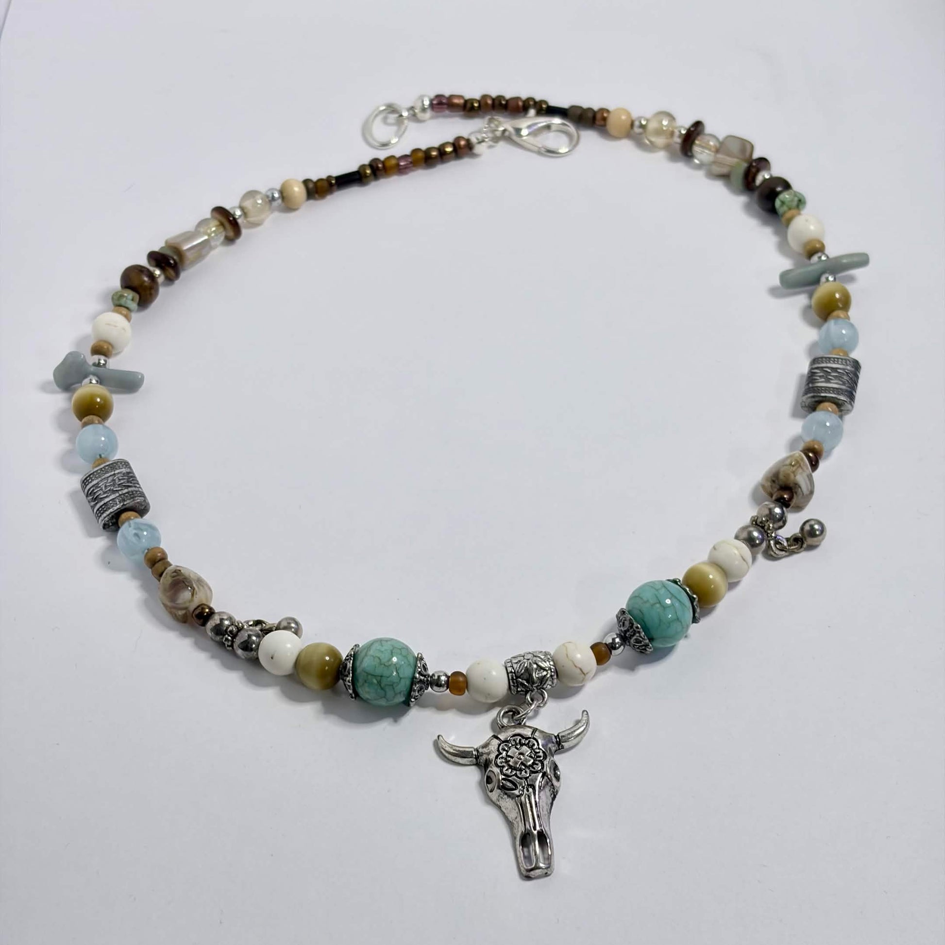 A beaded necklace with a silver-toned steer skull pendant, featuring light blue, white, brown, and gray beads, displayed on a white surface.