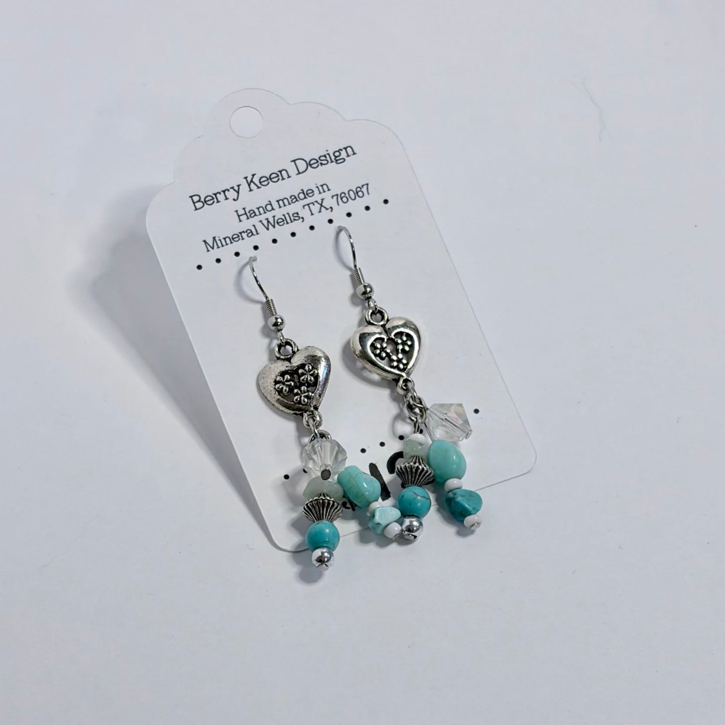 A pair of dangling earrings on a white display card. The earrings have silver-toned metal charms and beads with turquoise blue accents, photographed on a white surface.