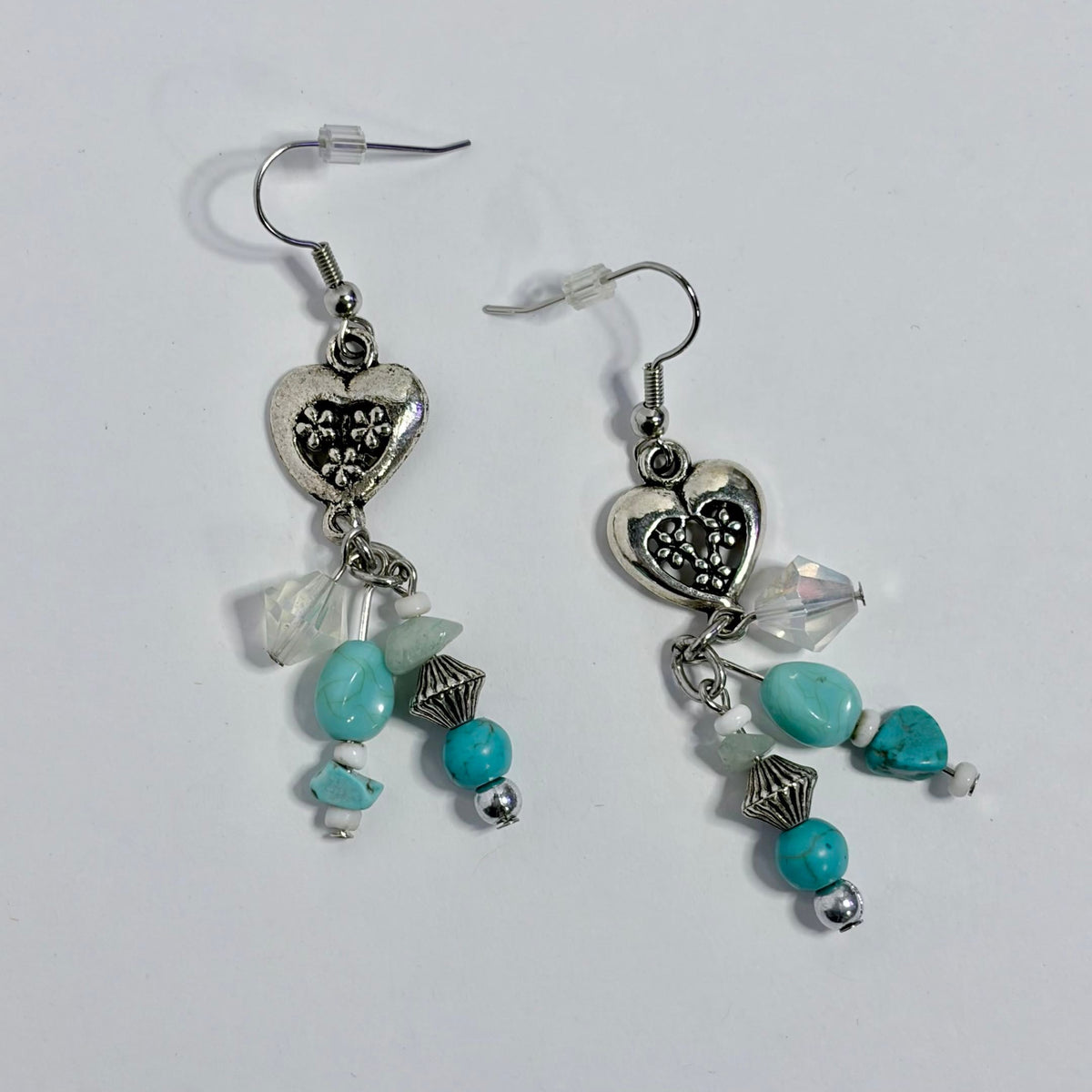 A pair of silver-toned dangle earrings with heart charms, turquoise beads, and clear faceted beads, displayed on a plain light background.