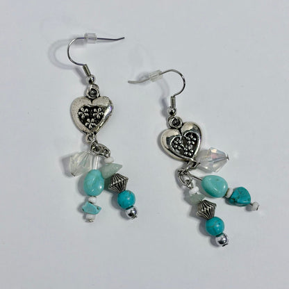 A pair of silver-toned dangle earrings with heart charms, turquoise beads, and clear faceted beads, displayed on a plain light background.