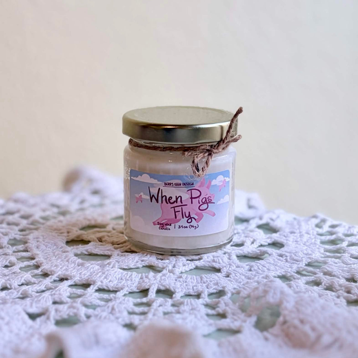 A small glass jar candle with a gold metal lid sits on a white crocheted doily on a pale green tabletop against a beige wall. The label reads 'When Pigs Fly".
