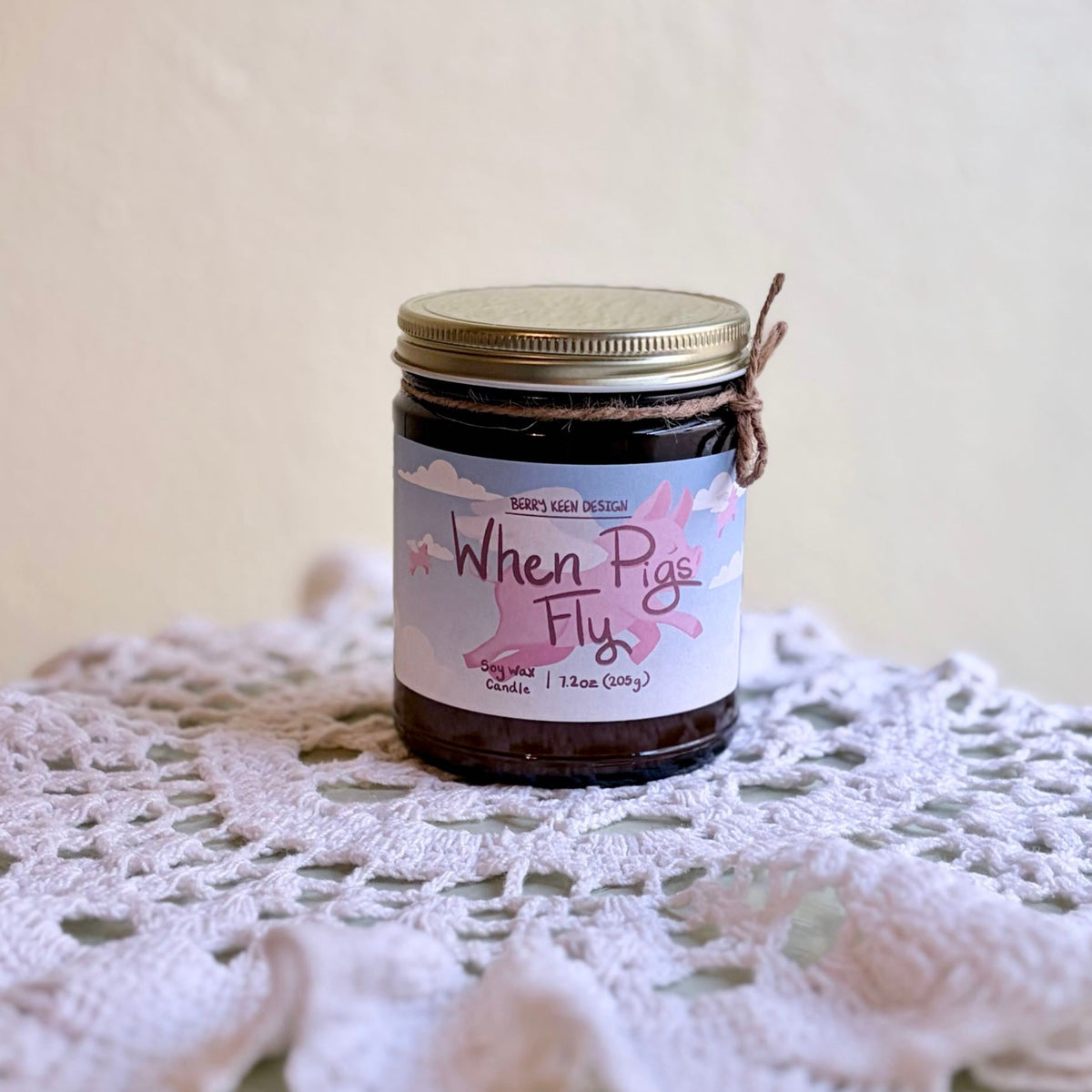 Amber candle jar "When Pigs Fly" on a doily with a beige background